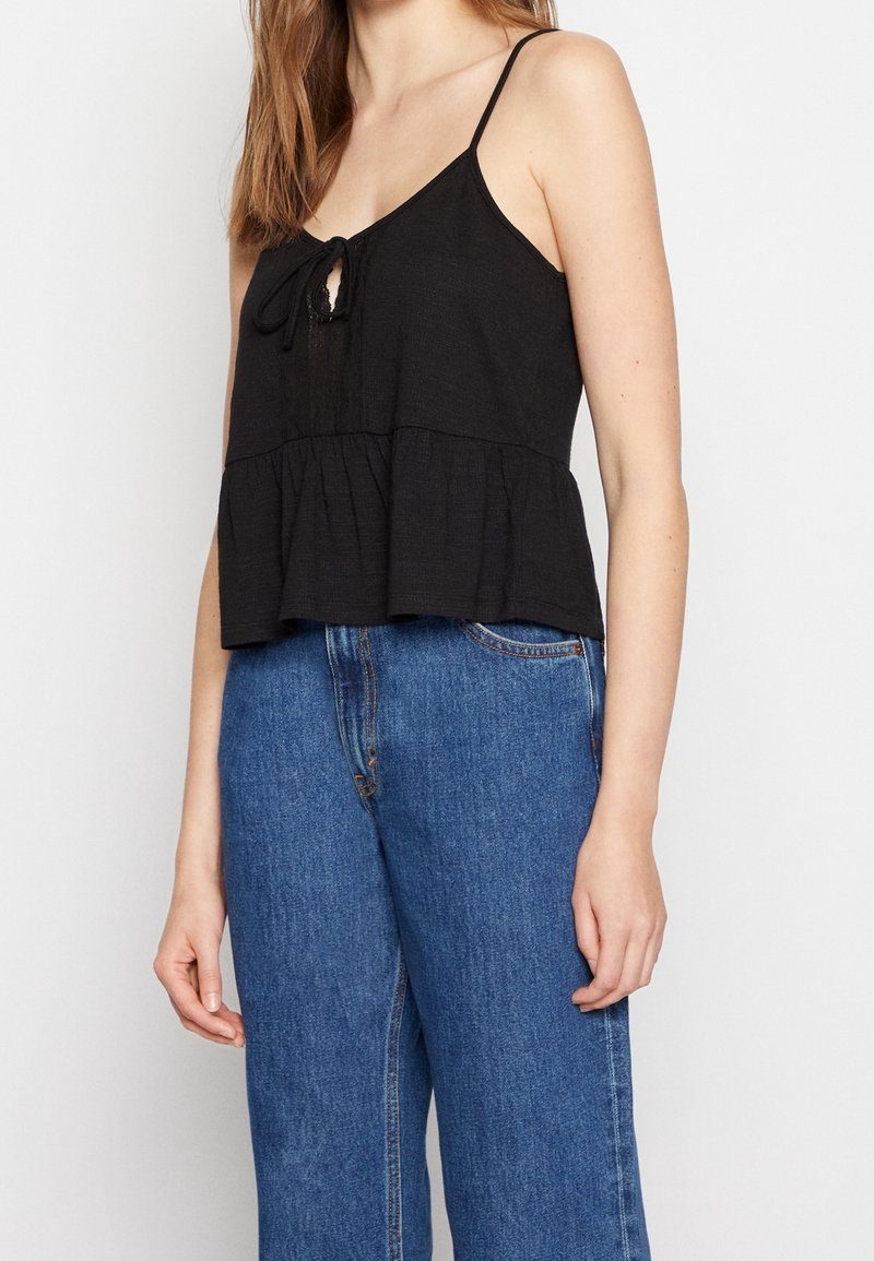 Haut en lin noir avec bretelles spaghetti ajustables, présentant un détail de nouage à l'avant et un style peplum évasé, associé à un jean bleu taille haute.