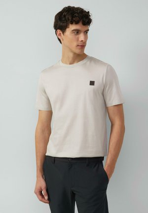 Light beige short-sleeve t-shirt made of soft fabric, featuring a small dark logo patch on the left chest. Paired with black pants.