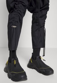 Schwarze Slip-On-Sneaker mit strukturierten Gummisohlen, die gelbe Akzente und eine Zuglasche aufweisen. Getragen mit schwarzen, schmal zulaufenden Hosen und Socken.