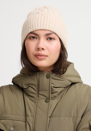 Berretto beige a maglia con texture a costine e un motivo a treccia nella parte superiore, indossato con un giubbotto imbottito verde oliva con cappuccio e bottoni a scatto.