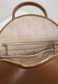 Brown leather backpack with a circular shape, featuring a beige interior, zippered pocket, and DKNY logo pattern on the lining.