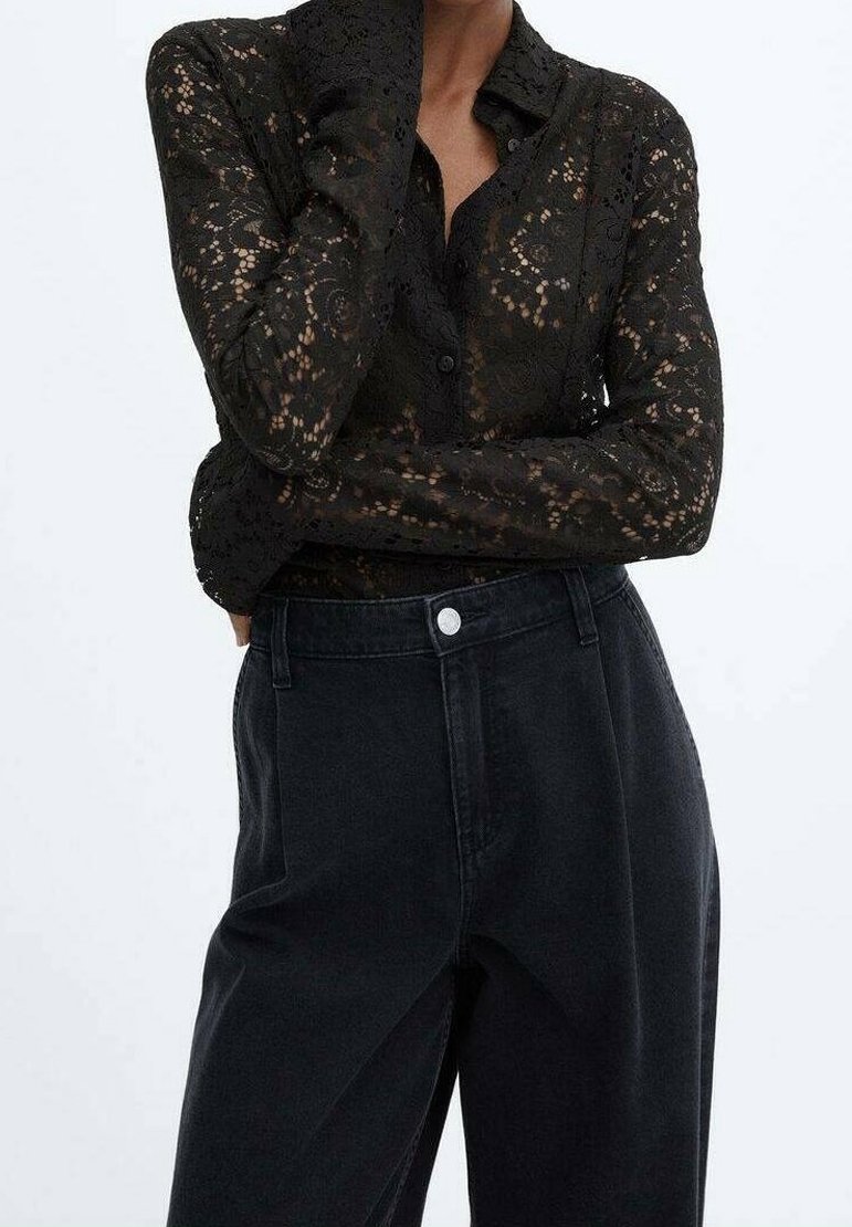 Chemise noire en dentelle à manches longues avec des motifs floraux portée avec un pantalon noir taille haute, bras croisés et main touchant le visage.