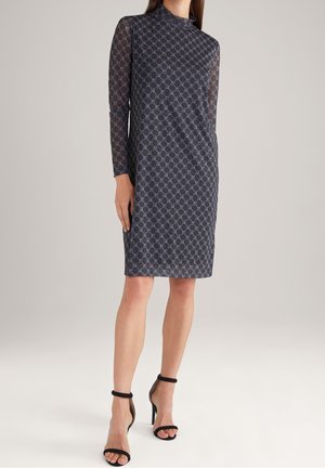 Femme portant une robe sombre à motifs, au genou, à manches longues, et des sandales noires à talons hauts avec bride à la cheville, debout sur un fond uni.