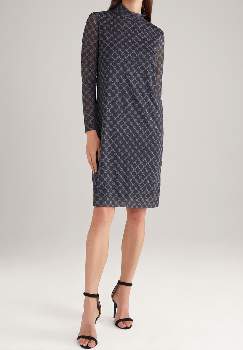 Femme portant une robe sombre à motifs, au genou, à manches longues, et des sandales noires à talons hauts avec bride à la cheville, debout sur un fond uni.