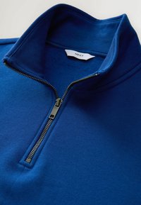 Suéter azul con cuello con cremallera, hecho de tela texturizada. Presenta una cremallera de metal y una etiqueta que dice "NEXT". Acabado suave en general.