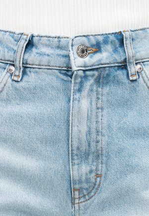 Lyseblå denimjeans i taljeområdet med metal knap og bæltestropper, båret med en hvid ribbet top, der er stoppet ind.