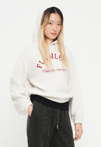 Κρεμ φούτερ με κουκούλα και κόκκινο κείμενο που αναγράφει "Ανώτερη Τελειότητα" και μαύρες λεπτομέρειες, συνδυασμένο με σκούρα joggers με σχέδιο.