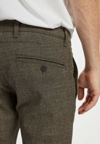 Brūnas raibās bikses ar teksturētu audumu, kurām ir viena aizmugurējā kabata ar melnu pogu. Jostasvietas daļa ir pieguļoša un plakana.