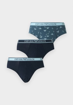 Izmos férfi áll mezítláb, teal színű Emporio Armani alsónadrágot visel világoskék derékrésszel és logómintával, balra néz, kezét a háta mögé helyezve.