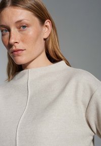 Beige, leichter Pullover mit hohem Ausschnitt, subtiler Textur und einer Naht detail, die vorne entlangläuft; kurze Ärmel und lockere Passform.