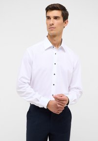 Witte lange mouwen blouse met zwarte knopen en contrasterende details aan de binnenkraag, gecombineerd met donkere broek. Soepele stoftextuur.