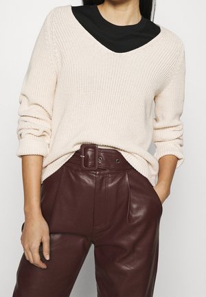 Femme portant un pull en maille crème par-dessus un top noir avec un pantalon en cuir bordeaux taille haute et une large ceinture, mains dans les poches.