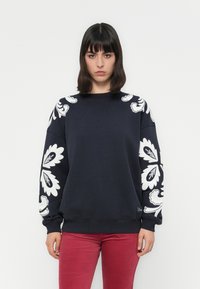 Sudadera azul marino con estampados florales blancos en las mangas, puños acanalados y escote. Presenta un corte relajado y una textura suave. Combinada con pantalones rojos.