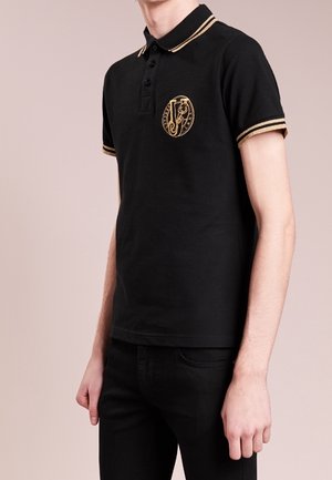 Homme portant un polo noir avec des finitions dorées sur le col et les manches, avec un emblème doré orné sur la poitrine gauche.