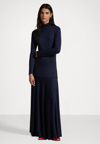 Vestido de manga larga en navy con cuello alto, que presenta una falda maxi fluida y una textura suave. Acabado con calzado rojo.