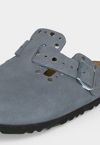 Μπλε σουέντ παπούτσι slip-on με διάτρητες μεταλλικές οπές και ρυθμιζόμενο λουράκι με αγκράφα πάνω σε μαύρη ραβδωτή σόλα.