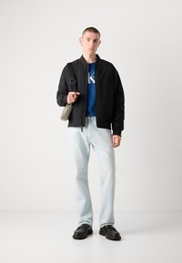 Jachetă bomber neagră, tricou grafic albastru, blugi deschiși, pantofi negri și geantă de umăr gri. Modelul stă în fața unui fundal alb.