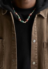 Man in een zwarte hoodie onder een bruine knopenjack met een kraalketting met groene, rode, witte en beige kralen.