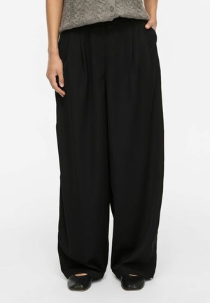 Personne portant un pantalon noir ample à jambes larges, un haut sans manches gris texturé et des chaussures plates noires, debout les mains près des poches.