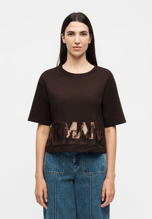 Kobieta o długich ciemnych włosach, ubrana w luźny czarny krótki T-shirt z napisem "ARMANI" oraz niebieskie jeansy z wysokim stanem, stojąca na białym tle.
