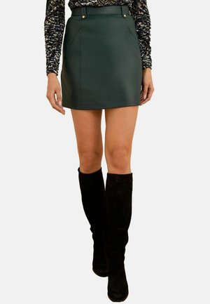 Donna vestita con una mini gonna verde scuro, un top a maniche lunghe con stampa leopardata e stivali neri al ginocchio, in piedi contro uno sfondo bianco.