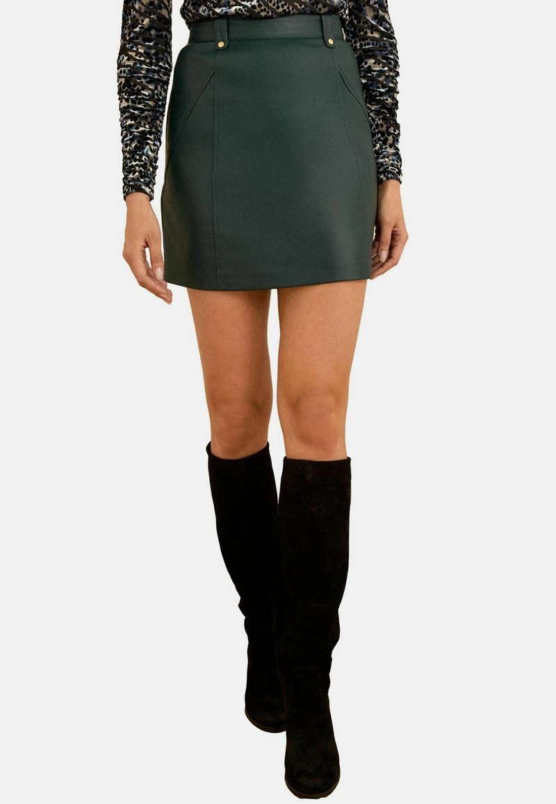 Femme portant une mini-jupe vert foncé, un haut à manches longues à imprimé léopard et des bottes montantes noires se tenant devant un fond blanc.