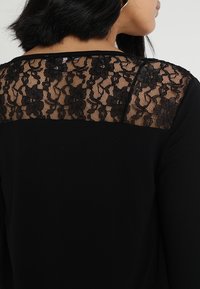 Czarny top z przezroczystym koronkowym karczkiem na plecach z kwiatowymi wzorami. Główny materiał jest miękki i jednolity, co kontrastuje ze szczegółami koronki.