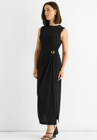 Black sleeveless dress with a twisted design at the waist, featuring a gold hardware accent. Smooth texture and floor-length cut.