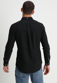 Hombre vestido con una camisa negra de manga larga con botones blancos, de pie de espaldas, combinada con jeans de mezclilla azules, sobre un fondo simple.