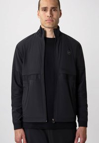 Black jacket with a high collar, zip front, mesh accents on sleeves, and a hexagonal logo on the chest. Smooth texture, sporty design.