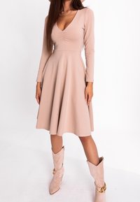 Robe longue à manches longues couleur nude avec un corsage ajusté, un décolleté en V et une jupe évasée. Fabriquée en tissu doux, avec un léger froncé subtil au niveau de la poitrine.