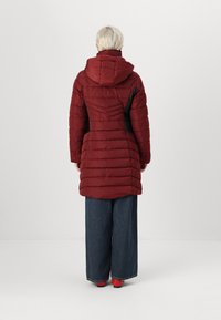 Manteau matelassé rouge avec une capuche, présentant une texture en matelassage horizontal et des panneaux latéraux noirs. Porté sur un pantalon en denim à jambes larges et des chaussures rouges.