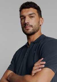 Donkergrijs polo-shirt met een kraag en een sluiting met twee knopen, vervaardigd uit textuurstof en met ribgebreide mouwen, gedragen door een persoon met gekruiste armen.