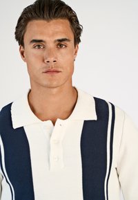 Νεαρός άνδρας με κοντά σκούρα μαλλιά που φοράει ένα πλεκτό πόλο μπλουζάκι με λευκές και ναυτικές ρίγες, κοιτάζοντας απευθείας την κάμερα σε απλό φόντο.