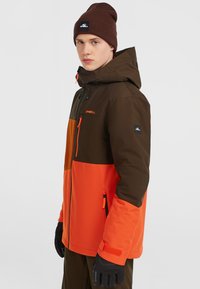 Kleurblokkering jas in bruin, oranje en donkergrijs met een ritszak op de borst, verstelbare capuchon en elastische manchetten. Gecombineerd met een bruine beanie.