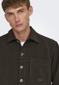 Camisa marrón de botones con cuello, que presenta botones de metal, un bolsillo en el pecho y costuras en contraste sobre una tela texturizada.