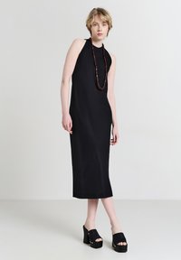 Schwarzes-neckholder Kleid aus glattem Stoff mit einer einfachen Silhouette. Kombiniert mit klobigen schwarzen Plateauschuhen und einer Perlenkette.