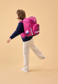 Roze kinderrugzak met meerdere zakken, banden en reflecterende accenten; bedrukte zijpatches; model draagt gestreepte broek en sneakers.