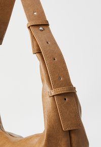 Detail einer braunen Ledertasche mit einem dicken, verstellbaren Tragegurt, der mit Nähten und Metallbeschlägen versehen ist. Glatte Oberfläche mit einer weichen, lässigen Form.