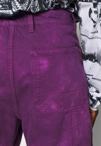 Pantalons cargo violets avec une surface légèrement texturée, comportant une poche arrière et des détails de couture contrastés. Portés avec une chemise à motifs.