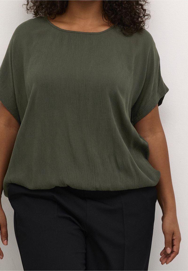Oliven grøn, løstsiddende top med korte ærmer og rund halsudskæring. Tekstureret stof, parret med mørke, skræddersyede bukser.