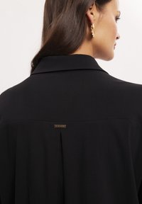 Czarna koszula z usztywnionym kołnierzykiem, centralnym zakładką na plecach oraz małą metalową metką blisko dekoltu.