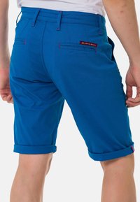 Blaue Shorts aus Baumwolle mit umgeschlagenen Säumen, kontrastierenden roten Nähten und zwei Gesäßtaschen mit einem Logo-Patch.