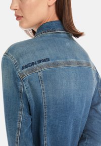 Denim jacket in light blue, featuring a collar, back yoke, and "MRGIAN SPORTS" embroidered in dark blue on the left shoulder area.