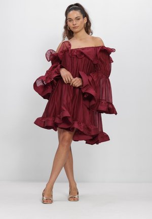 Vrouw staand in een bordeauxrode off-shoulder jurk met ruches aan mouwen en zoom, met gouden open teen hakken tegen een effen achtergrond.
