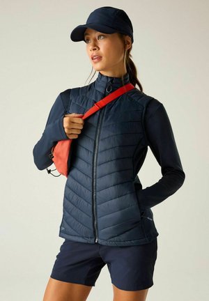 Woman wearing a navy jacket, shorts, and cap, holding a red sling bag strap, looking to the side against a plain background.