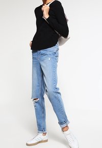 Maglione nero a maniche lunghe, jeans azzurri strappati con strappi, sneaker bianche con dettagli grigi e verdi, e uno zaino grigio.