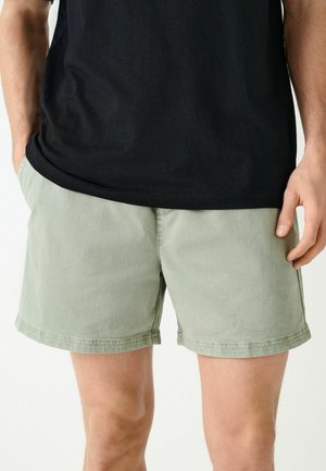 Férfi világoszöld hétköznapi rövidnadrágban és fekete pólóban, oldalt laza kéztartással áll egyszínű háttér előtt.