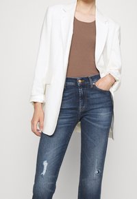 Blazer bianco sopra una canotta marrone a coste, abbinata a jeans blu strappati con un ginocchio strappato e una vestibilità attillata.