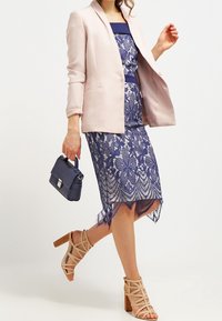 Marinblå spetsklänning med scallopdetaljer i fållen, i kombination med en ljusrosa blazer och hudfärgade strappy klackar. En liten marinblå handväska fulländar looken.